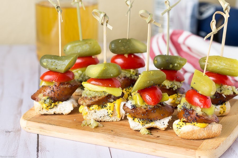 pan tostado, rebanada gruesa de salchicha ahumada, condimento dulce, cebolla blanca, mostaza amarilla, lanza de pepinillo, semillas de sésamo, todo en un bocado en un pincho