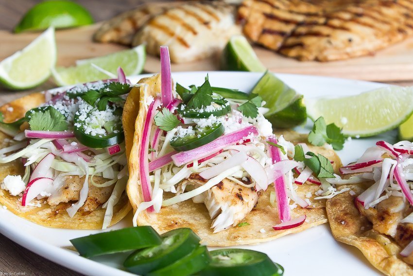 Tacos de pescado a la parrilla marinados con cerveza, estos tacos clásicos están cubiertos con repollo rallado, rábanos, cebollas rojas en escabeche, rodajas de jalapeño y queso cojita.