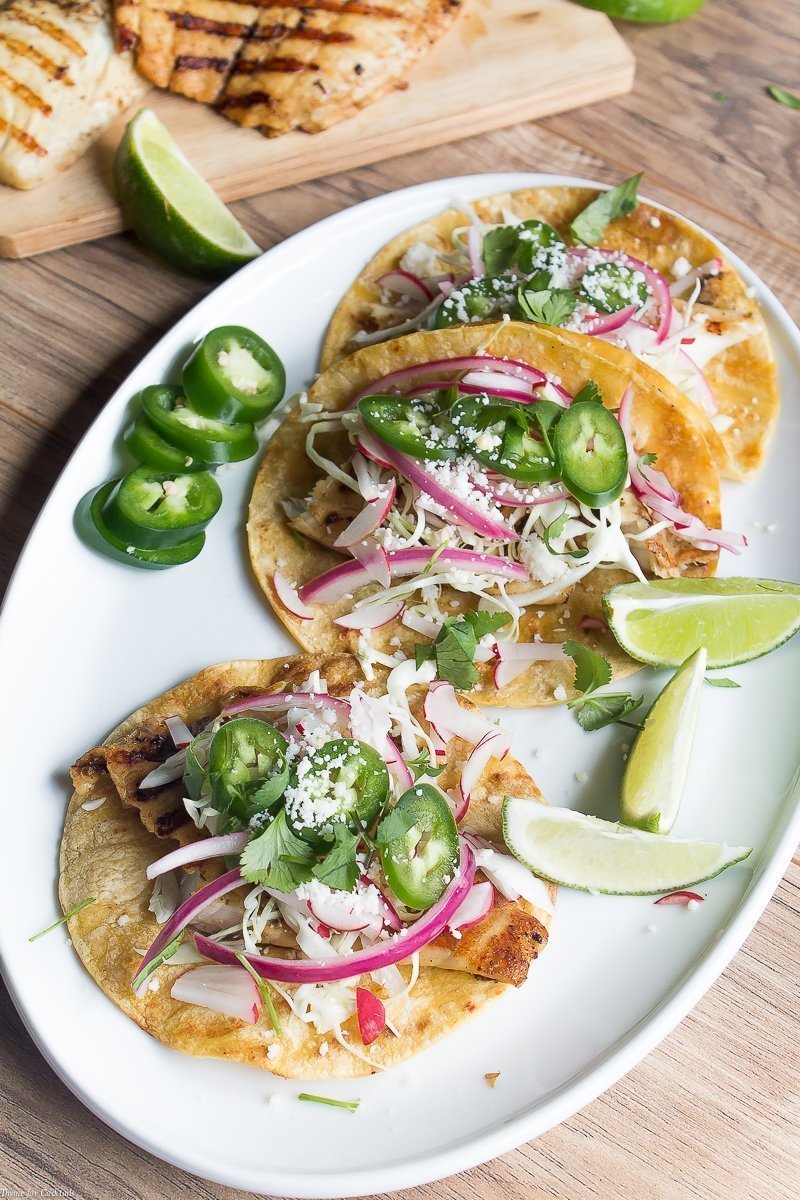 Tacos de pescado a la parrilla marinados con cerveza, estos tacos clásicos están cubiertos con repollo rallado, rábanos, cebollas rojas en escabeche, rodajas de jalapeño y queso cojita.
