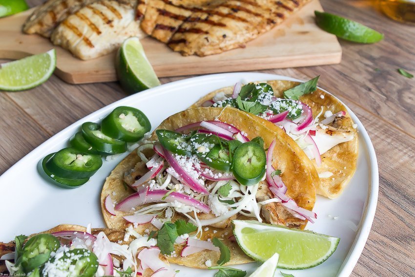 Tacos de pescado a la parrilla marinados con cerveza, estos tacos clásicos están cubiertos con repollo rallado, rábanos, cebollas rojas en escabeche, rodajas de jalapeño y queso cojita.