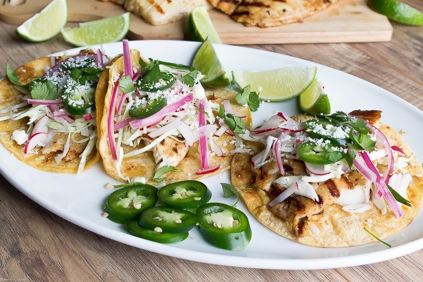 Tacos de pescado a la parrilla marinados con cerveza, estos tacos clásicos están cubiertos con repollo rallado, rábanos, cebollas rojas en escabeche, rodajas de jalapeño y queso cojita.