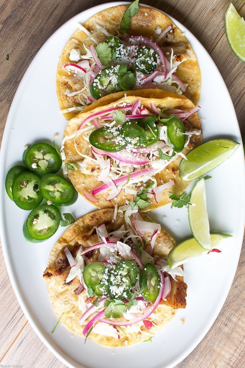 Tacos de pescado a la parrilla marinados con cerveza, estos tacos clásicos están cubiertos con repollo rallado, rábanos, cebollas rojas en escabeche, rodajas de jalapeño y queso cojita.